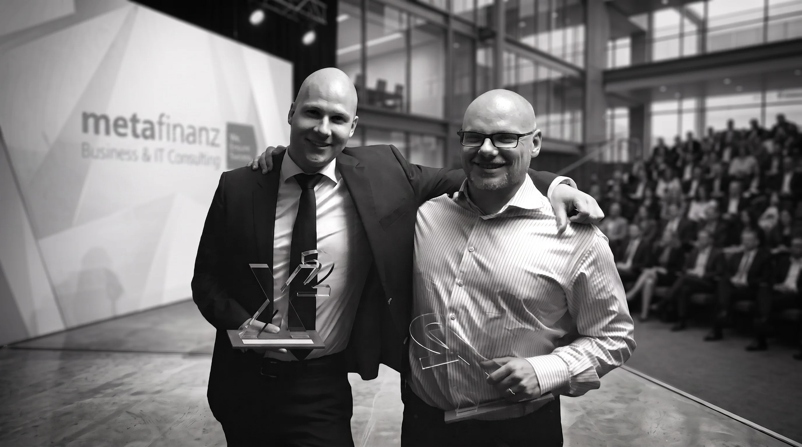 Felix Schmidt (left) and a colleague on stage at the metafinanz X-cellence Award ceremony, both holding clear acrylic trophies, with the metafinanz logo and the audience in the background