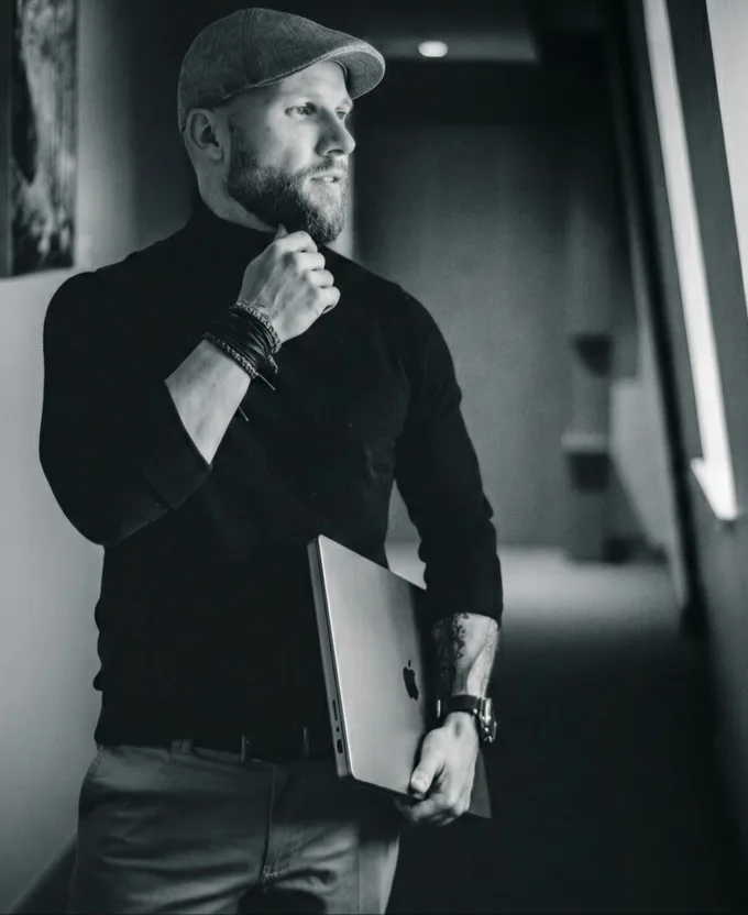 Black-and-white portrait of Felix Schmidt wearing a flat cap and black turtleneck, holding a MacBook under his arm, looking thoughtfully to the side in a hallway